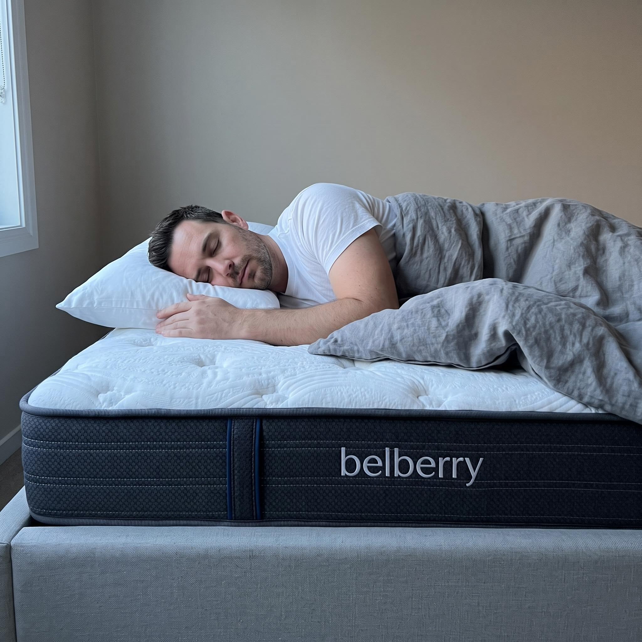 A man sleeping soundly on a Belberry Support mattress with a white pillow and gray bedding. The mattress has a dark navy blue side panel with the brand name 'belberry' printed in white.